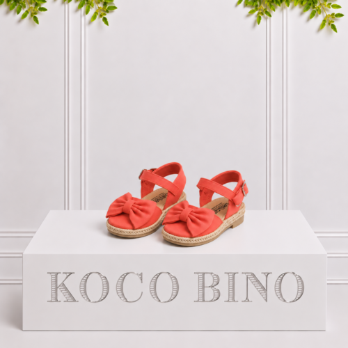 Coral girls’ espadrille sandals with large bows and ankle straps displayed on a white Koco Bino pedestal with leafy greenery.
