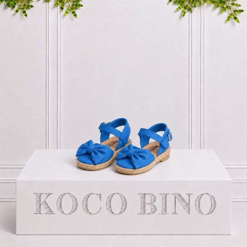 Ocean blue girls’ espadrille sandals with large bows and ankle straps displayed on a white Koco Bino pedestal with leafy greenery.