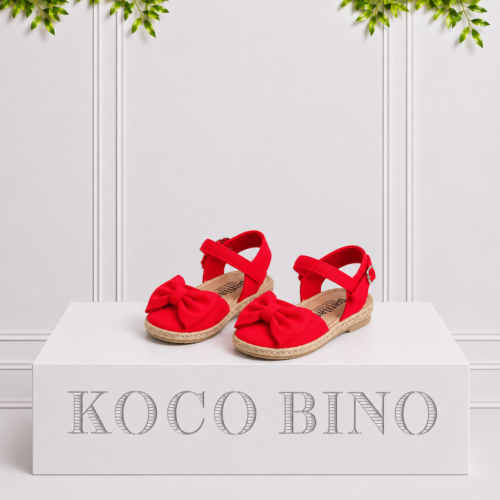 Red girls’ espadrille sandals with large bows and ankle straps displayed on a white Koco Bino pedestal with leafy greenery.