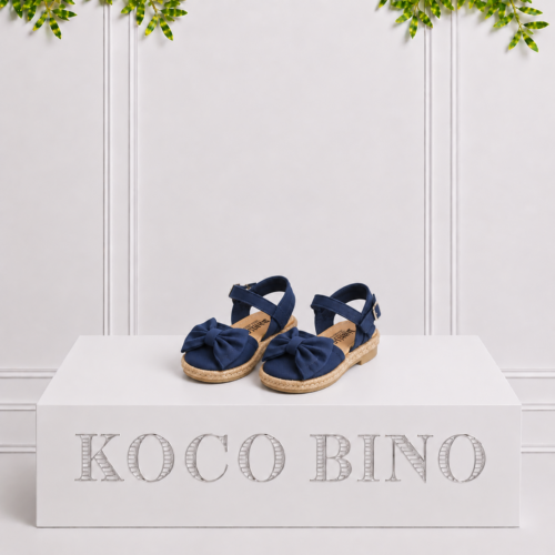 Navy girls’ espadrille sandals with large bows and ankle straps displayed on a white Koco Bino pedestal with leafy greenery.