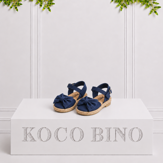 Navy girls’ espadrille sandals with large bows and ankle straps displayed on a white Koco Bino pedestal with leafy greenery.
