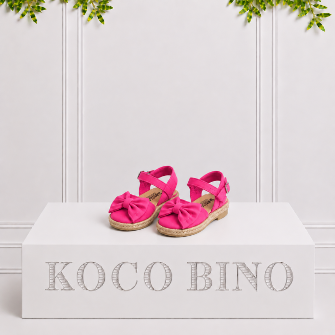 Fuchsia girls’ espadrille sandals with bows and ankle straps displayed together on a white Koco Bino pedestal with leafy greenery