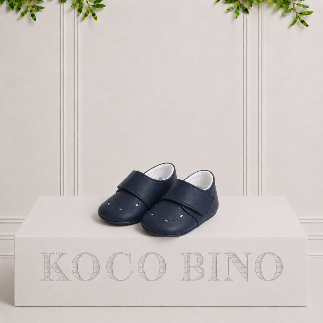 Kai navy leather baby shoes with a classic strap fastening, displayed on a Koco Bino podium in an elegant studio setting.