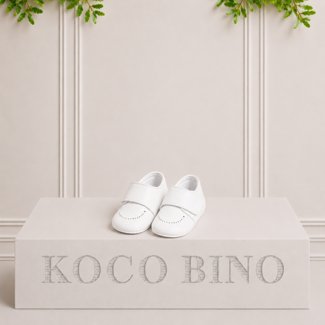 Kai white leather baby shoes with a classic strap fastening, displayed on a Koco Bino podium in an elegant studio setting.
