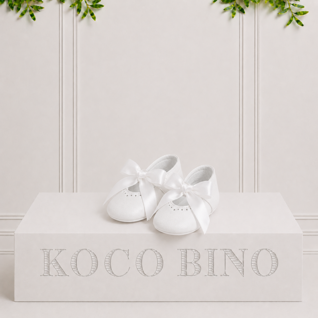 Amy white leather baby shoes with satin bows displayed on a Koco Bino podium in a clean studio setting with elegant décor.