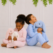 Boy and girl wearing matching blue and pink cotton pyjamas sitting back to back with plush bunnies in a cosy Koco Bino studio scene.