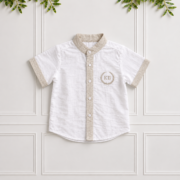 White boys' linen shirt with stone collar, cuffs and button placket, featuring a KB laurel embroidery on a cream panelled wall.