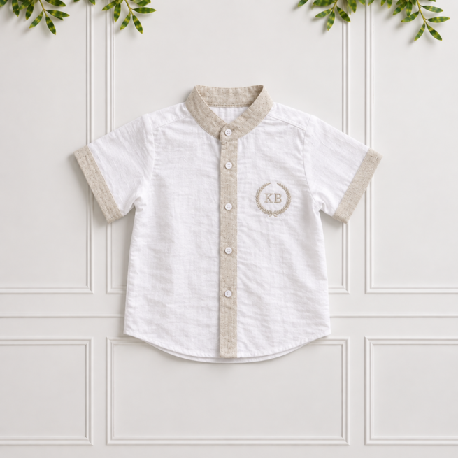 White boys' linen shirt with stone collar, cuffs and button placket, featuring a KB laurel embroidery on a cream panelled wall.