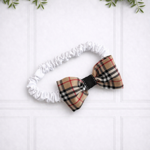 Baby girl bow headband featuring a stone tartan bow on a soft white ruched band, beautifully presented against a white panelled background.