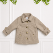 Beige children's coat with button front, classic collar and rolled cuffs, displayed on a white paneled wall with green foliage above, front view.