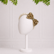 Elegant cream satin girls' headband with a sparkling gold sequin bow, displayed on a white mannequin stand with leafy accents above.