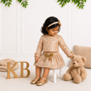 Little girl wearing a gold lace top and matching skirt with a velvet bow, gold shoes and headband, seated beside a teddy bear in an elegant studio setting.