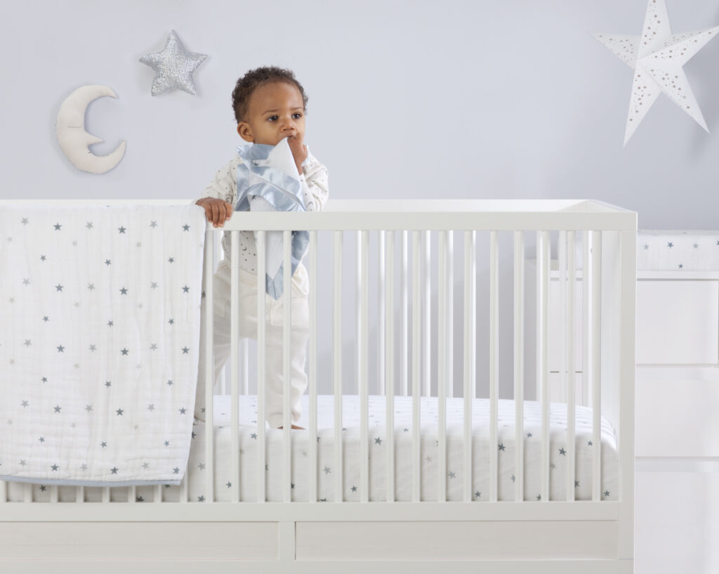 Baby standing in a white cot with an Aden + Anais muslin blanket featuring grey stars, creating a soft, stylish and cosy nursery scene.