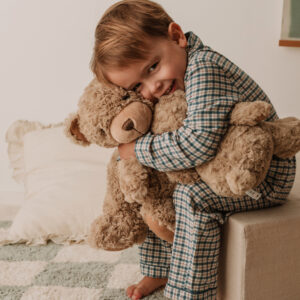 Young child cuddling a soft teddy bear while sitting in a cosy nursery, wearing checked pyjamas and smiling warmly in a comforting setting.