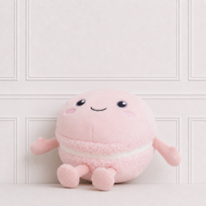 Soft pink macaron plush toy with embroidered smiling face, rosy cheeks and little arms and legs, displayed on a neutral background for décor.