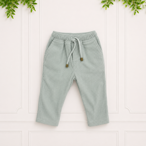 Sage green baby corduroy track pants with drawstring waist and gold cord ends, displayed flat on a white paneled background with green foliage above.