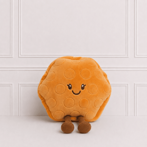 Cute waffle plush toy with embroidered smiling face and dangling legs, sitting on a neutral studio background with elegant wall panelling in soft light.
