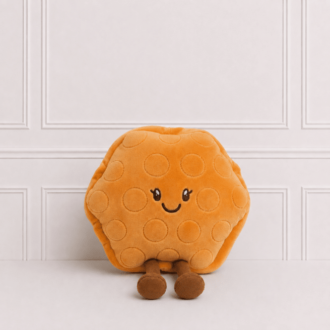 Cute waffle plush toy with embroidered smiling face and dangling legs, sitting on a neutral studio background with elegant wall panelling in soft light.