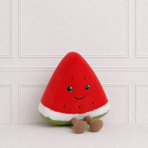 Soft watermelon slice plush toy with embroidered smile and dangling legs, sitting on a neutral studio background with elegant wall panelling.