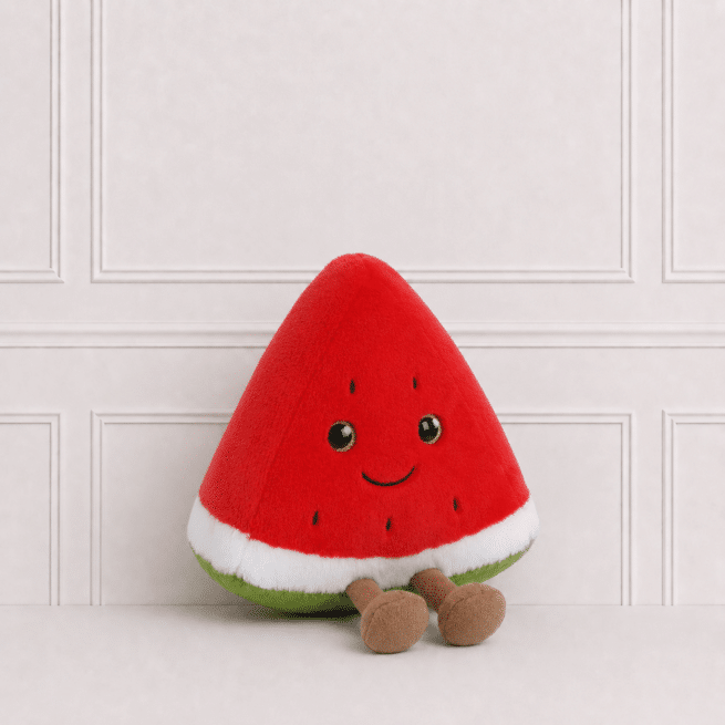 Soft watermelon slice plush toy with embroidered smile and dangling legs, sitting on a neutral studio background with elegant wall panelling.