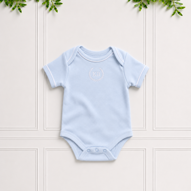Blue baby bodyvest in soft cotton interlock with envelope neckline, white KB embroidery and white piping, displayed on a classic Koco Bino background.