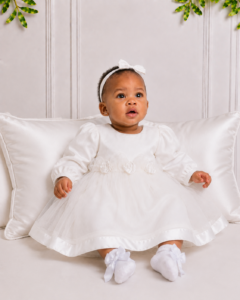 Koco Bino baby girl christening dress with white bow headband and satin socks, seated on silk cushions in an elegant South African studio setting.