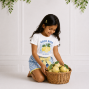 Young girl wearing a white lemon print T-shirt and blue gingham shorts with a yellow bow in a bright studio.