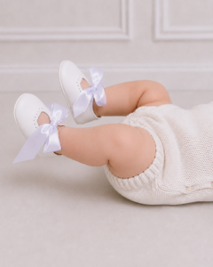 Baby girl shoes with white satin bows on a baby lying down, showcasing soft leather first walking shoes for special occasions and christenings.