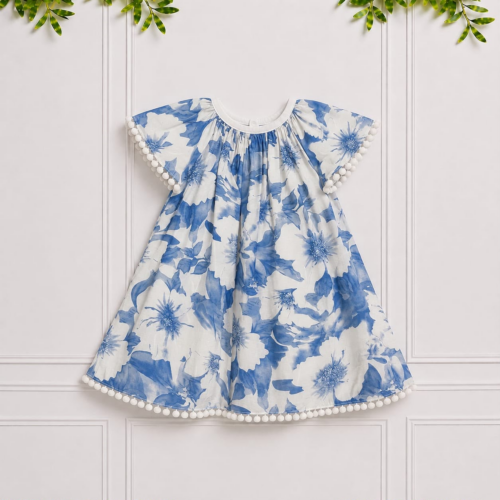 Emily girls' blue and white floral dress with white pom-pom trim and bow, displayed against an elegant white paneled studio background.