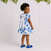 Young girl wearing a blue and white floral dress with white back bow and pom-pom trim, smiling over her shoulder in a studio setting with greenery.