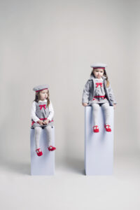 Two girls model a Koco Bino grey winter outfit with matching berets, coats and red shoes in a minimalist studio photoshoot.