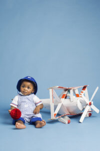 Koco Bino baby boy wearing a blue striped romper and blue sun hat, sitting beside a vintage toy aeroplane in a playful studio photoshoot.