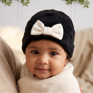Baby girl wearing a black velvet beanie with a white bow, wrapped in a cosy cream knitted blanket in a warm, elegant setting.