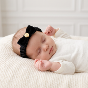 Sleeping baby wearing a delicate black velvet headband with a gold detail, resting peacefully in a cosy white outfit.