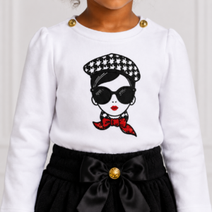 Close-up of a baby girl wearing a white long-sleeve top with a stylish graphic, gold shoulder buttons and a black bow skirt.