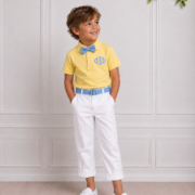 Young boy wearing a yellow polo, blue bow tie and belt, white cuffed trousers and white loafers, posing in a bright studio.