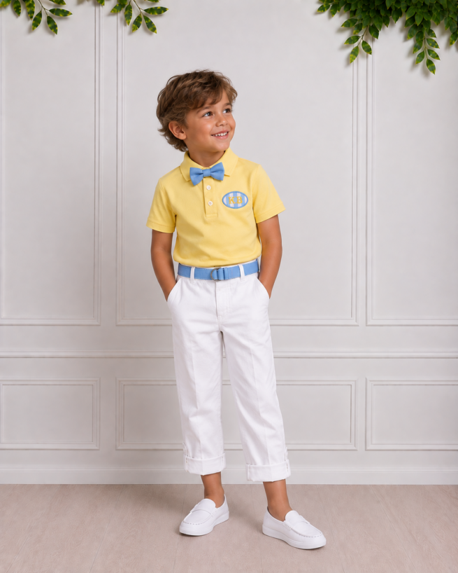 Young boy wearing a yellow polo, blue bow tie and belt, white cuffed trousers and white loafers, posing in a bright studio.