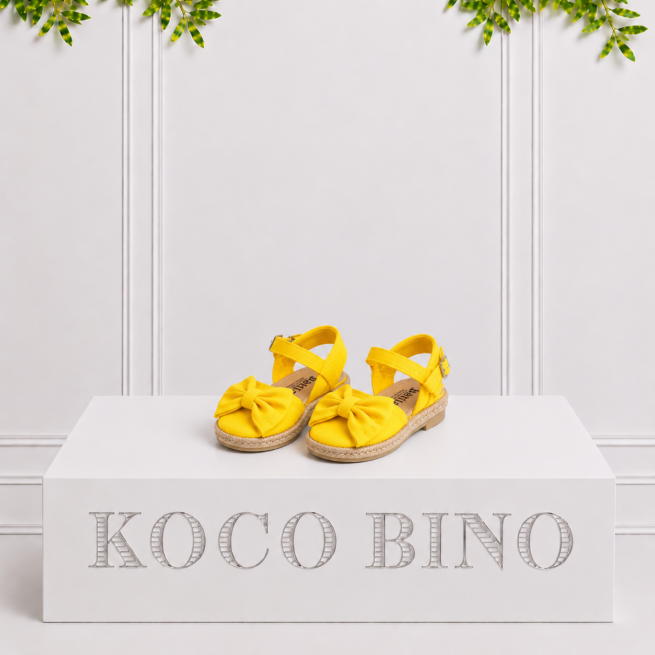Bright yellow girls’ espadrille sandals with bows and ankle straps displayed together on a white Koco Bino pedestal against a leafy backdrop for kids