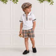 Young boy wearing boys check shorts with a matching white polo, driver cap and black moccasins in a cream studio, styled in classic tartan.