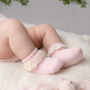 Baby wearing soft pink socks with delicate lace trim, resting on a cosy cream blanket in a soft, elegant nursery setting.