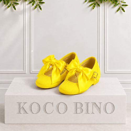 Girls yellow pumps with oversized bows and buckle straps displayed on a white Koco Bino pedestal