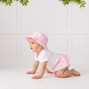 Baby girl wearing pink bloomers with a white top and matching sun hat, posing on a soft studio background with a charming, elegant look.