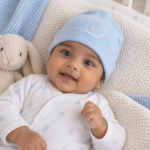 South Asian baby boy wearing a soft blue knitted beanie with KB embroidery, smiling while lying on cosy cream bedding beside a plush bunny.
