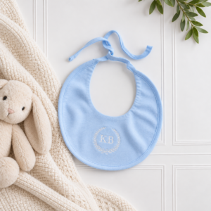 Pale blue baby bib with delicate KB embroidery, styled on a white panelled backdrop with a cosy cream knitted blanket and soft bunny.