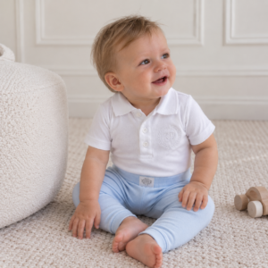 Smiling baby boy wearing pale blue leggings and a white collared top, sitting barefoot on a cream rug beside a cosy boucle ottoman.