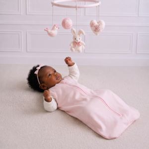 Baby girl wearing a soft pink sleeping bag with a white long-sleeve top, lying beneath a charming nursery mobile.