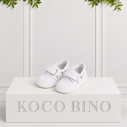 White children's boat shoes with Velcro straps, stitched detailing and textured soles displayed on a Koco Bino branded pedestal against a cream backdrop.