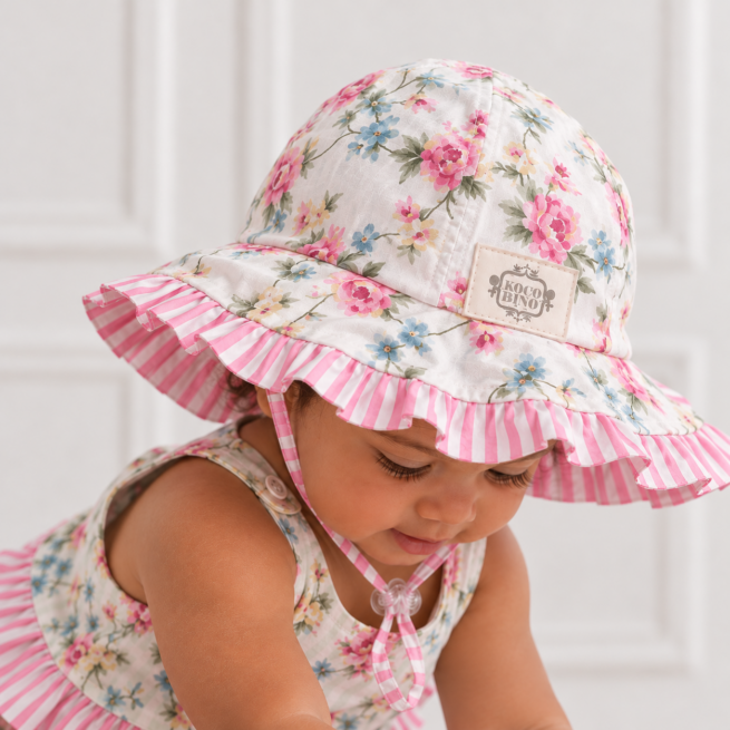 Baby girl wearing a floral sunhat with pink striped frill and toggle tie, looking down in a close-up showing the hat’s natural creases and details.