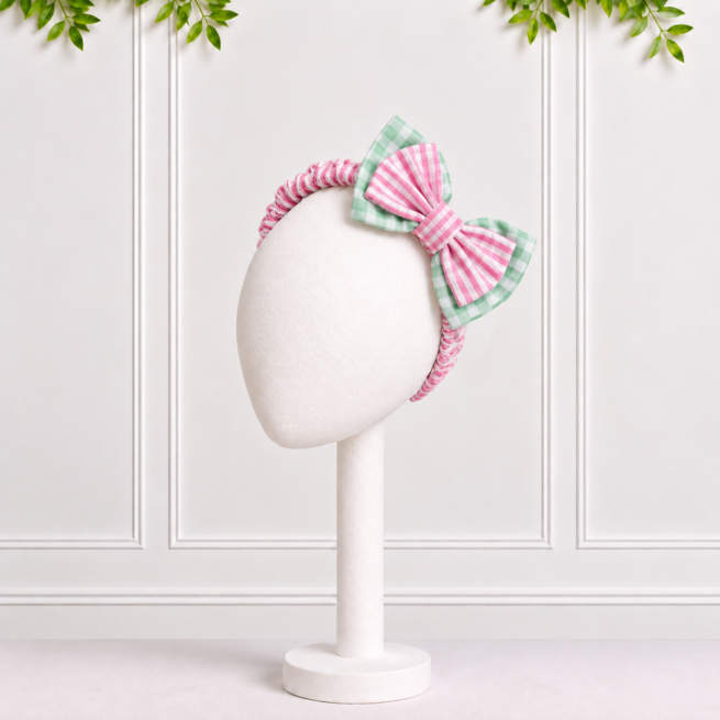 Baby girl pink and green gingham bow headband with pink striped centre, displayed on a white mannequin in a studio setting.