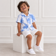 Indian boy laughing joyfully on a white cube, wearing a blue palm print shirt, white shorts and white shoes, in a bright studio.